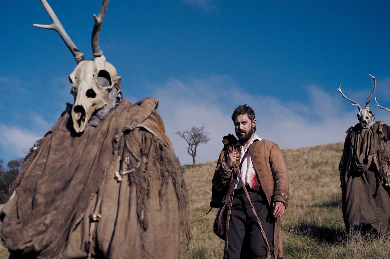 a man with a beard is standing in a field with a deer skull on his head