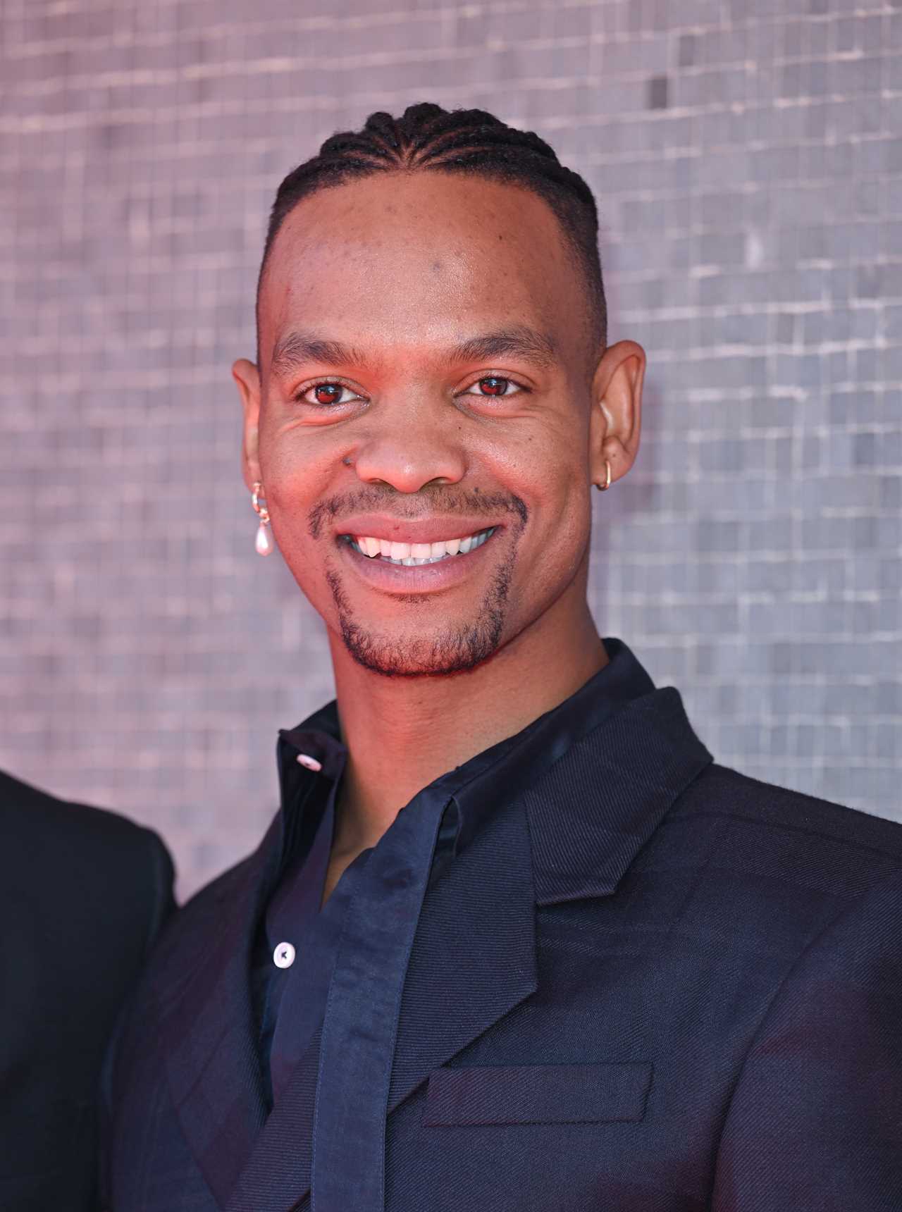 a man with braided hair and earrings smiles for the camera